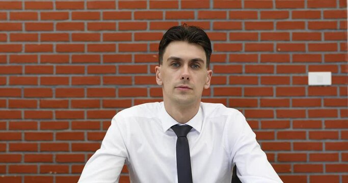 Young adult businessman in casual white shirt sitting in front of red brick wall listen to speaker in online conference and nod head with agreed and then raise hand for ask question.