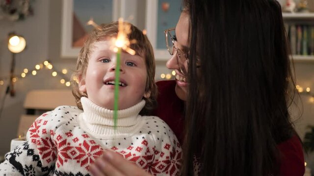 Christmas lights, sparklers. Happy smiling woman and child 2-3 years old look at sparkler, Christmas garland in background. Close-up and slow motion