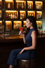 Nightlife concept a gorgeous girl in a blue collared dress looking confident sitting beside the bar sipping the pink cocktail