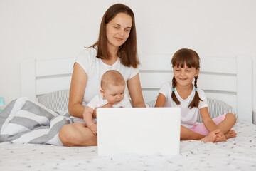 Indoor shot of happy positive female with dark hair sitting on bed with her children, with two little girls, trying to work from hone, freelance and parenting,