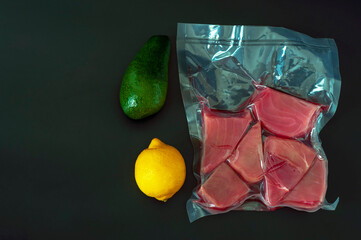 Raw tuna steak in a vacuum package, avocado and lemon on a black background. The concept of storing food, healthy food and ingredients for cooking-salads, tartare and other dishes. copy space.