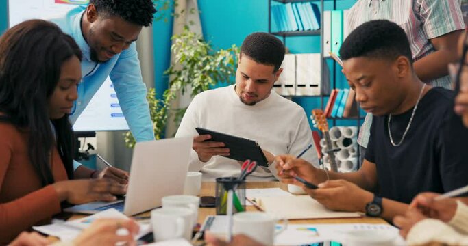 A group of students sit in a classroom at a table with books, notes, and a computer and work on a project for college. They are reviewing ideas on the tablet, jotting down notes, discussing
