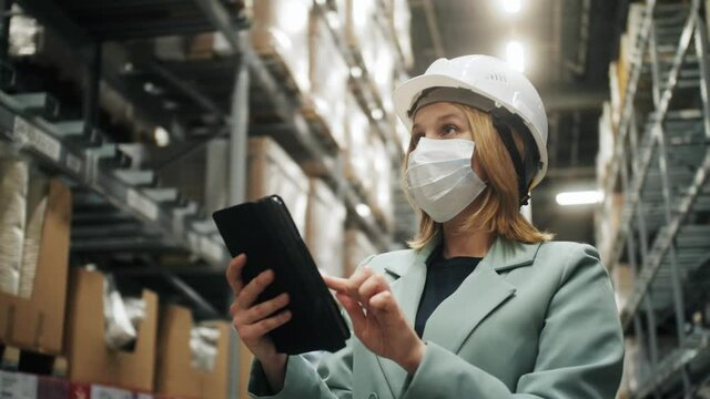 Female manager of warehouse wearing protective face mask and hard hat checking products stock in warehouse and inventory with tablet standing in retail warehouse full of shelves with goods.