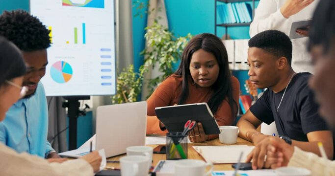 Dark-skinned friends work together in company on graphics for new design logo displaying inspirations on tablet discussing searching for ifnromation creating taking notes talking possible changes