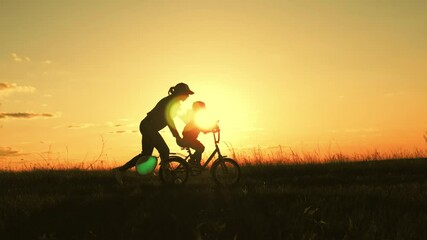 Silhouette of mother teaching little son to ride a bike at meadow during sunset. The kid turns the bike close-up. Caring young mother teaches her son to ride bicycle. Concept of friendly family.