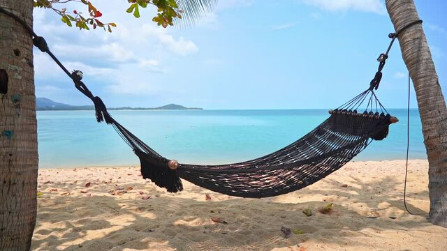 Empty cottom rope net hammok with spreader bars lashed between two palm trees on the sandy beach  in front of the turquoise sea at tropical island