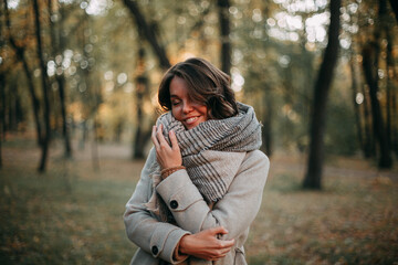 Smiling young brunette girl walking in an autumn park in a beige coat and a beige scarf