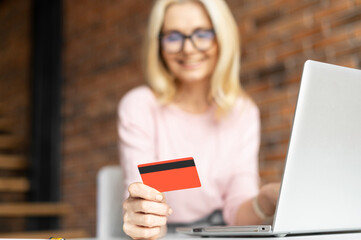 Selective focus at the debit card in hand of pretty mature businesswoman wearing glasses sitting at the desk using laptop for online payment from home, ordering food, tranfering money. E-banking
