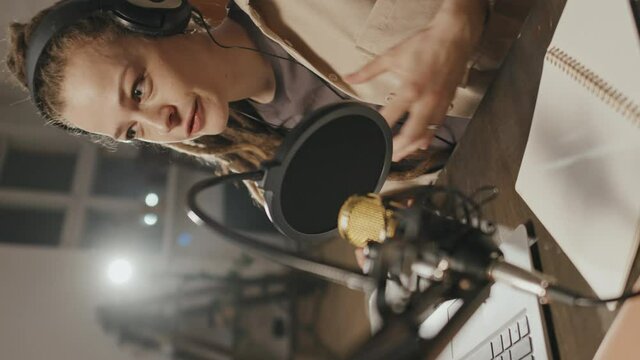 Handheld vertical shot of cheerful young woman with dreadlocks sitting at table at home or studio and talking into microphone. She is gesticulating and telling story on podcast