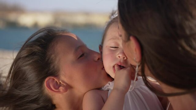 Close-up portrait of carefree pretty baby girl with cheerful sisters kissing toddler on cheeks in slow motion. Happy Caucasian child in sunshine outdoors with family on picturesque sea beach