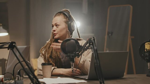 Tracking Of Cheerful Young Woman With Dreadlocks Sitting At Table In Dark Living Room And Talking Into Microphone While Having Conversation With Someone Off Camera And Recording Podcast At Home