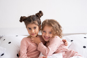 two beautiful diverse kids girls in pajamas hugging on bed in modern bright apartment