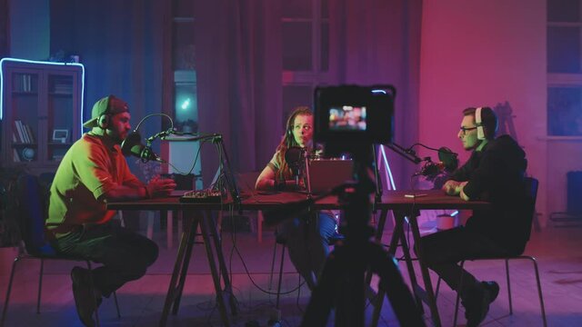 Tracking of young men and woman in headphones sitting at table in living room lit by neon string lights and having conversation while recording podcast. Video camera standing on tripod and filming