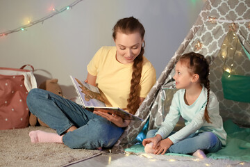 Young woman reading fairy-tale to her little daughter late in evening at home