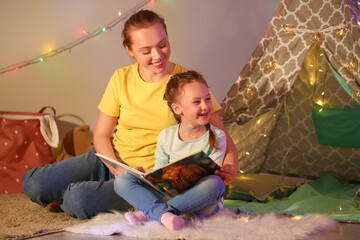 Young woman reading fairy-tale to her little daughter late in evening at home