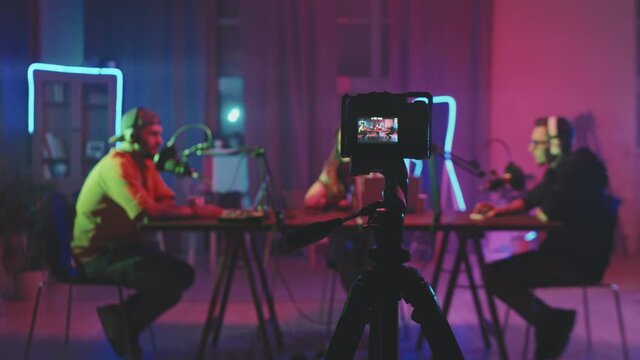 Tracking shot of video camera standing on tripod and filming three people sitting at table in living room lit by neon string lights and recording podcast