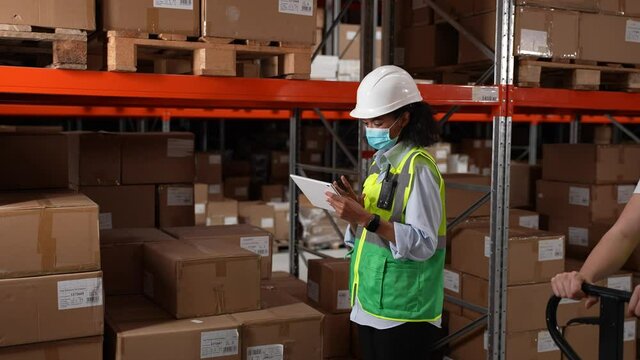 Mixed race female supervisor wearing face mask checking products stock in warehouse during coronavirus infection outbreak. Men workers passing by while inventory manager standing at rack with boxes