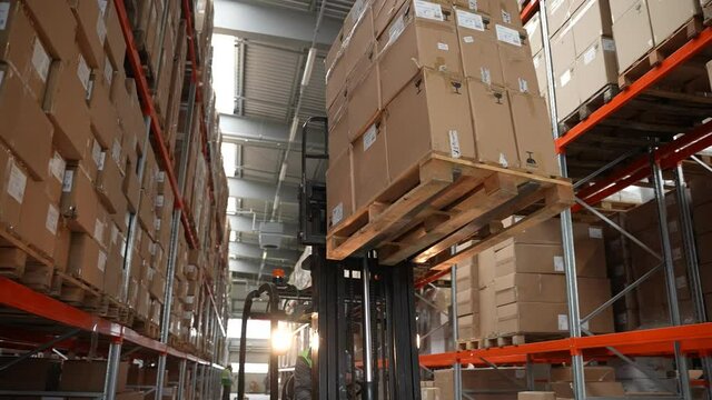 Storehouse worker moving large pallet of cardboard boxes unloaded from height. Male forklift driver in hard hat riding with goods between rows of racks in factory storage