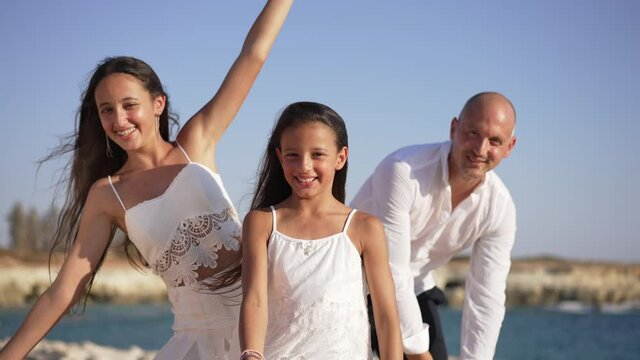 Joyful little girl smiling looking at camera as teenage sister and father bending to sides posing. Portrait of happy pretty Caucasian child standing with family on Mediterranean sea coast having fun