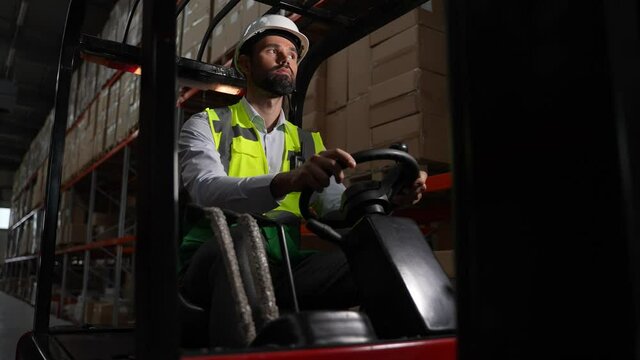 Confident bearded man in hard hat and vest riding loader backwards during work in storehouse. Serious warehouse employee driving forklift truck between racks with goods in cardboard boxes