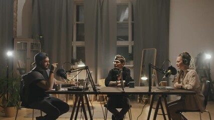 Tracking shot of three male and female podcast hosts sitting at table in dimly lit living room or studio and talking into microphones while recording show