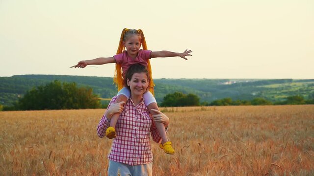 Mom and daughter on shoulders have fun in field. Family and nature in summer at sunset. Little girl and woman on vacation. Smile be happy. Friendship of two daughters can fly. Farmers play in field.