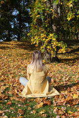 A teenage girl with long bleached hair sits on fallen leaves in the autumn forest