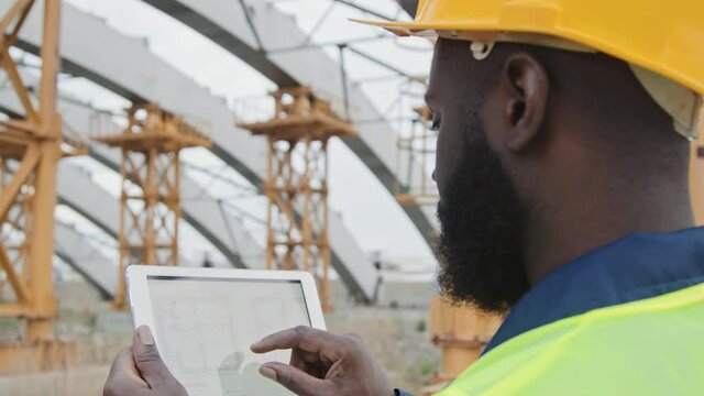 Tracking close up of black male engineer in safety vest and hard hat reading blueprint on tablet and looking at unfinished building with scaffolding at construction site