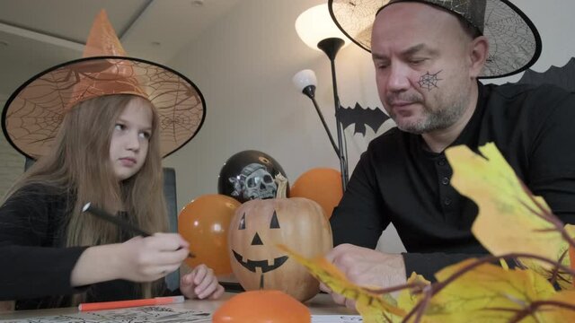 Child with father draw pictures on the theme of halloween in decorated room at home. Little girl and her dad smiling and having fun together, happy family preparing to celebrate the holiday.