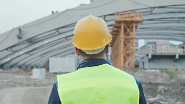 Slowmo tracking with rear view of male construction site foreman in safety vest and hard hat walking