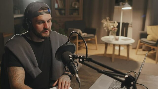 Handheld slowmo shot of man with beard sitting at table in cozy living room with dim lighting and talking into microphone while recording podcast at home