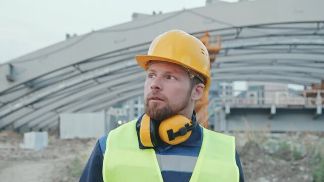 Slowmo tracking shot of bearded male construction site foreman in hard hat and safety vest looking around and walking outdoors at workplace
