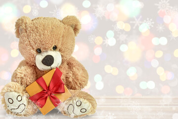 Teddy bear with a gift with a red ribbon sits on a light background with the addition of bokeh and snowflakes.  Christmas or New Year's decor.  place to add text.  card