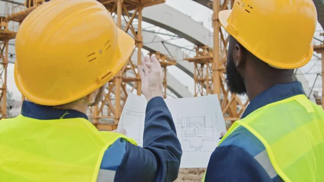 Slowmo tracking shot of Caucasian and African-American male construction workers in safety vests and hard hats discussing blueprint before unfinished building with scaffolding