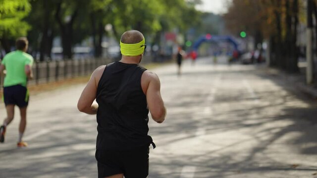 Slow motion muscular man wearing black sportswear and yellow headband approaching finish line at city marathon, other contestants on blurred background. Following shot runner at sports event