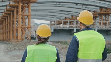 Tracking rear view of male and female construction site supervisors in safety vests and hard hats walking towards unfinished building or bridge with scaffolding and having conversation
