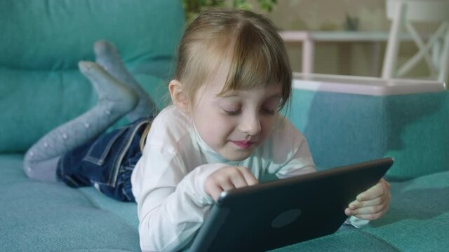 Preschooler Girl Playing Games Using Digital Tablet Lying On Sofa Alone. Curious Little Kid Girl Hold Pad Computer Watching Video At Home Children Education Or Tech Internet Addiction Concept