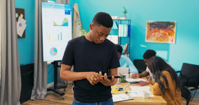 Proud, happy and smiling, dark-skinned erasmus student is group leader, supervising project work of colleagues in corporate office. The boy browses tablet raises head crosses arms on chest