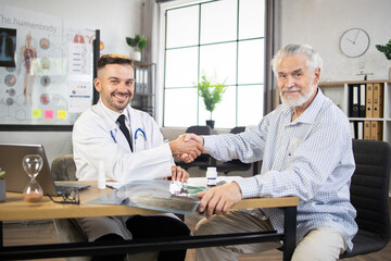 Smiling caucasian physician and retired male patient shaking hands while sitting at medical office....