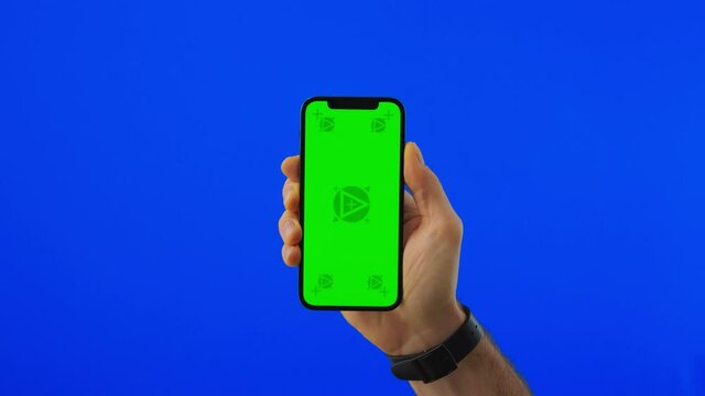 Hand with the Phone Appears from the Camera Side. Man Holding a Phone with Vertical Green Chroma Key Screen Isolated on Blue Background.