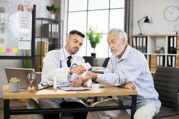 Obraz premium Caucasian medical worker in white lab coat measuring temperature of senior bearded man. Male doctor using modern thermometer during checkup of aged patient.