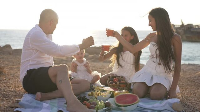 Happy Caucasian family clinking juice glass with baby girl in slow motion in sunshine on Mediterranean sea. Wide shot of relaxed father with Japanese Respect and Power tattoos and daughters on picnic