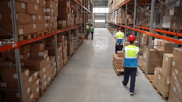 Aisle between racks with cardboard boxes in storehouse, back view of two workers following each other to place goods, two logistics managers communicating aside