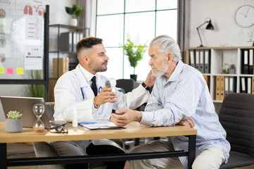Caucasian medical worker in white lab coat checking temperature of senior patient with modern infrared contactless thermometer. Examination of health condition at clinic.