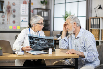 Caucasian mature woman in lab coat explaining aged man results of tomography. Competent female doctor holding x ray scan and talking with male patient. Concept of medical consultation.