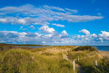 White cumulus clouds over the sand dunes with beach grasses and posted footpath. Blue ocean and lagoons in distance.