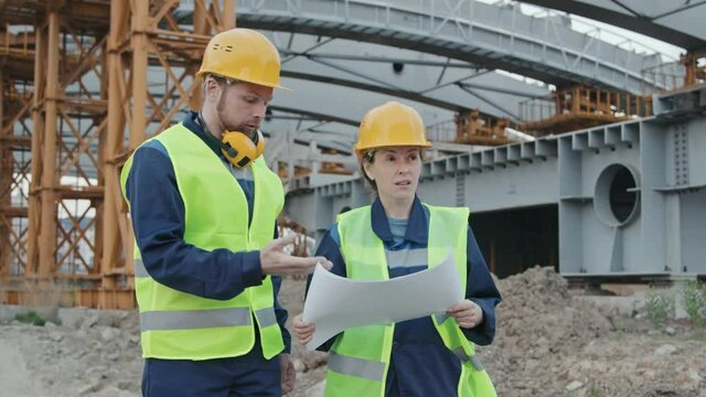 Tracking shot of male and female engineers in safety vest and hard hats discussing blueprint and walking on construction site with scaffolding
