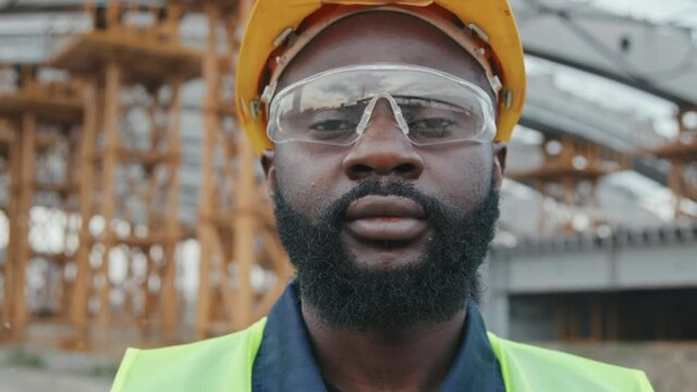 Tracking close up portrait shot of serious African-American foreman in hard hat and goggles standing at construction site and looking at camera