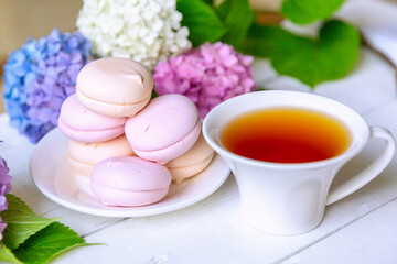 A white cup of classic tea with pink marshmallows surrounded by hydrangea flowers on a white wooden table.