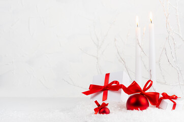 Christmas background with white burning candles, gift boxes, red decorations - glossy balls with satin ribbons in decorative soft light white winter forest with frosty branch and snow, copy space.